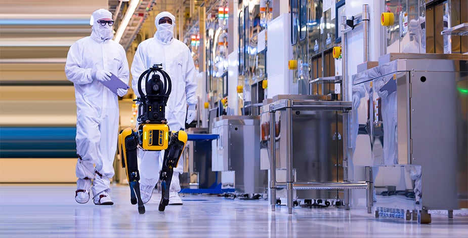 Two cleanroom-suited engineers walking a yellow quadruped robot through a fabrication facility