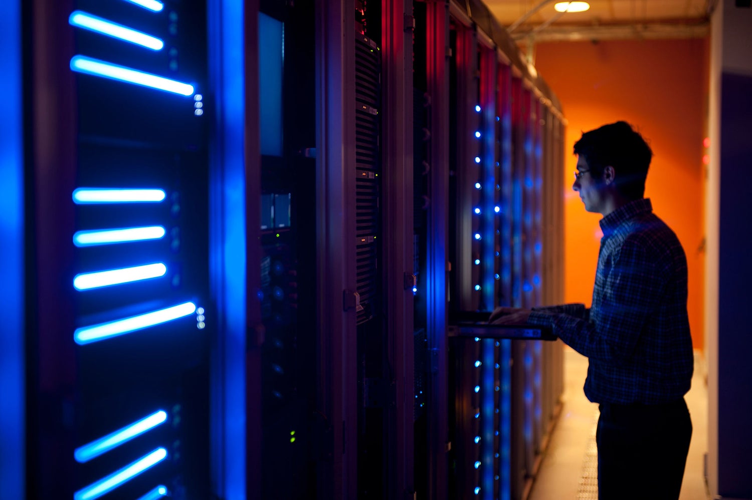 An engineer walking a dark data-center corridor lined with blue neon strips and warm amber depth